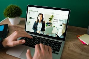 Person using a laptop with a website builder open on the screen. The interface features a smiling woman holding a laptop, with text reading “CREATE YOUR WEBSITE” and “START NOW.” A green wall and office supplies are visible in the background.